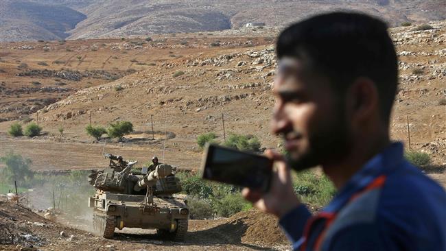 A Palestinian man holds a mobile phone as Israeli troops ride a howitzer during a military exercise in the Palestinian territories on November 2, 2016. (Photo by AFP)