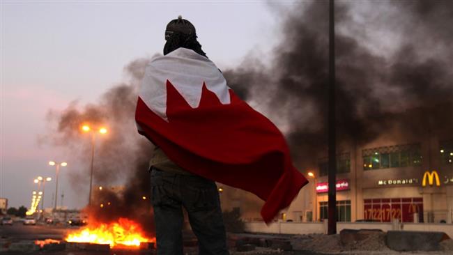In this file photo, a Bahraini anti-regime protester wearing a national flag watches for police as tires burn on a street in the western village of Malkiya, Bahrain. (Photo by AP)