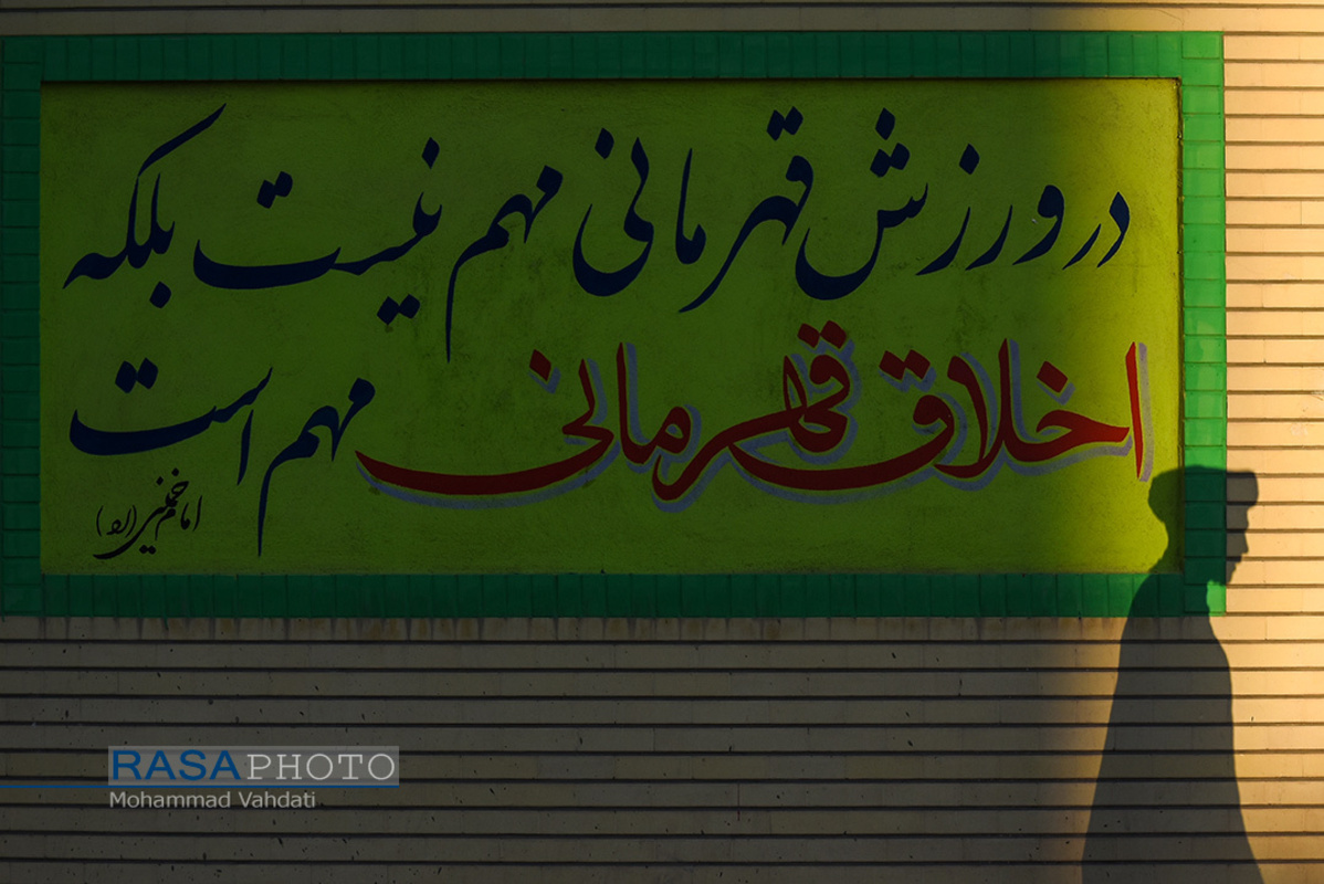 یک عکس یک کلام | طلبه ای که از کنار دیوار نوشته ای عبود میکند که بر روی آن کلامی حکیمانه از حضرت امام خمینی (ره) نوشته شده است که : در ورزش قهرمانی مهم نیست بلکه اخلاق قهرمانی مهم است.